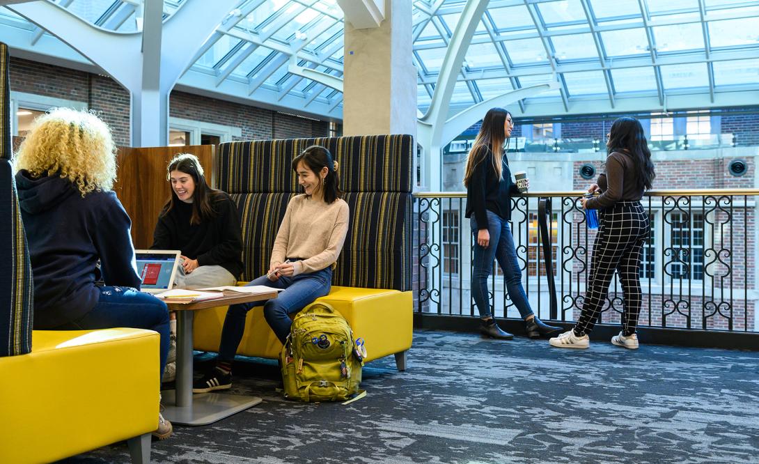 3 Students in the Idea Hub at the Michigan Union talking while sitting in a booth, two more students are standing at the rail